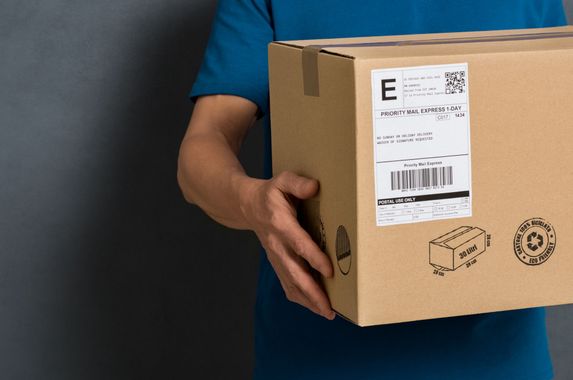 Person in blue shirt holding a cardboard package with a shipping label against a gray background.