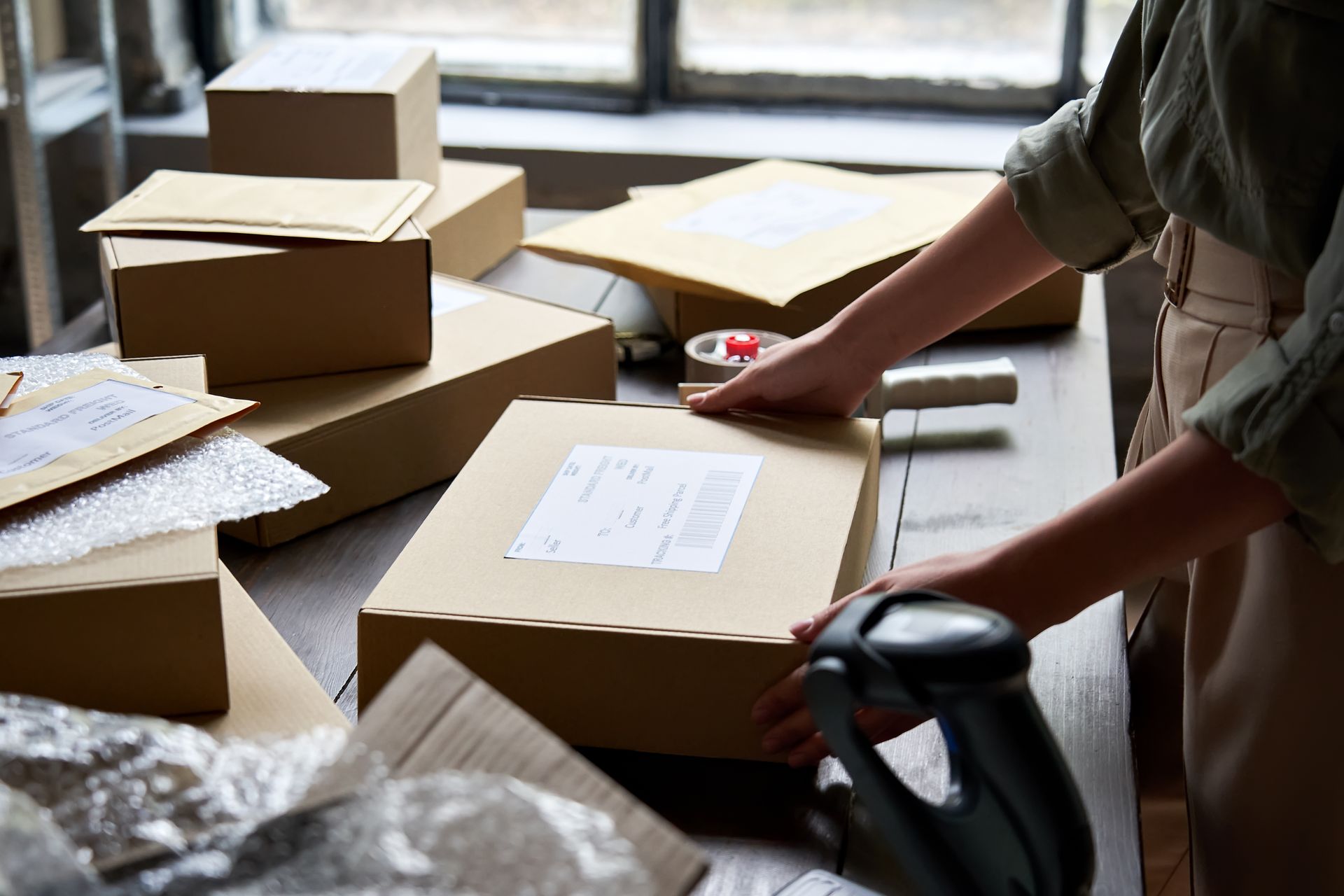 Person packing a cardboard box on a table, surrounded by other packages and shipping materials.