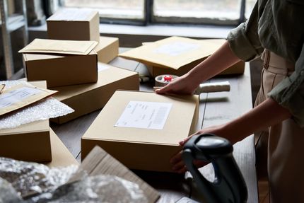 Person packing a cardboard box on a table, surrounded by other packages and shipping materials.