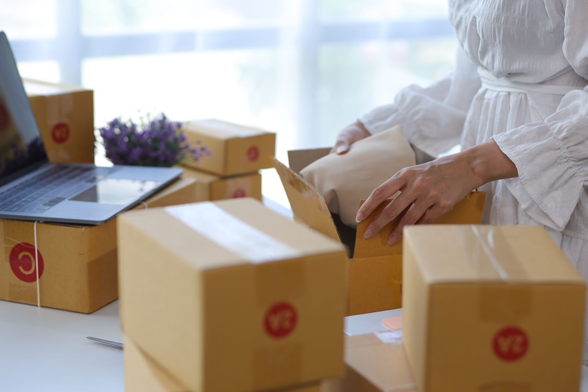 Woman packing packages for shipping; multiple cardboard boxes on a desk near a laptop.