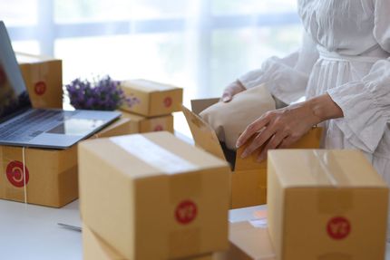 Woman packing packages for shipping; multiple cardboard boxes on a desk near a laptop.