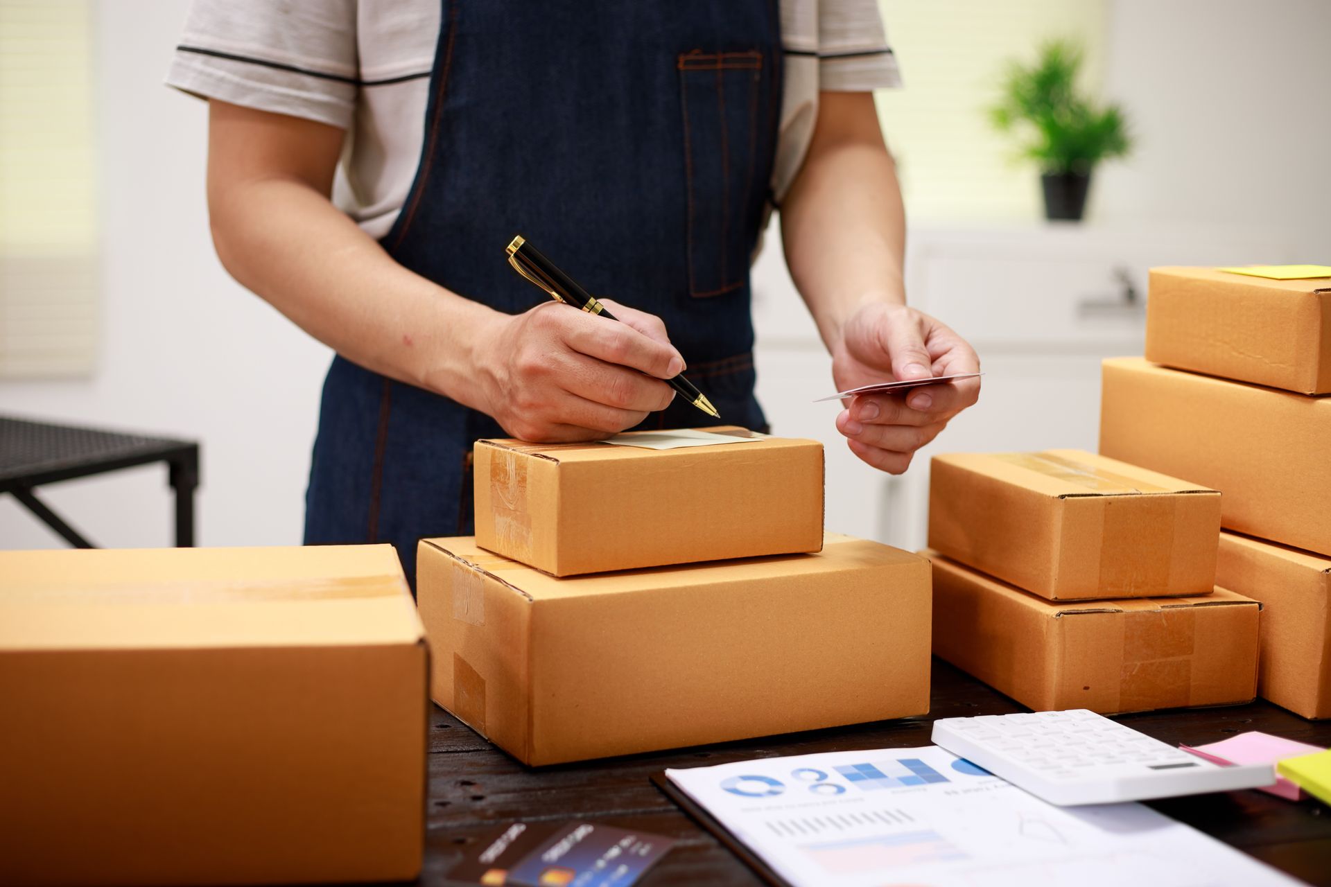 Person in apron signing shipping boxes, workspace with calculator and paperwork.