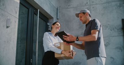 Delivery person handing a package to a woman at a doorway; the woman smiles and holds the box.
