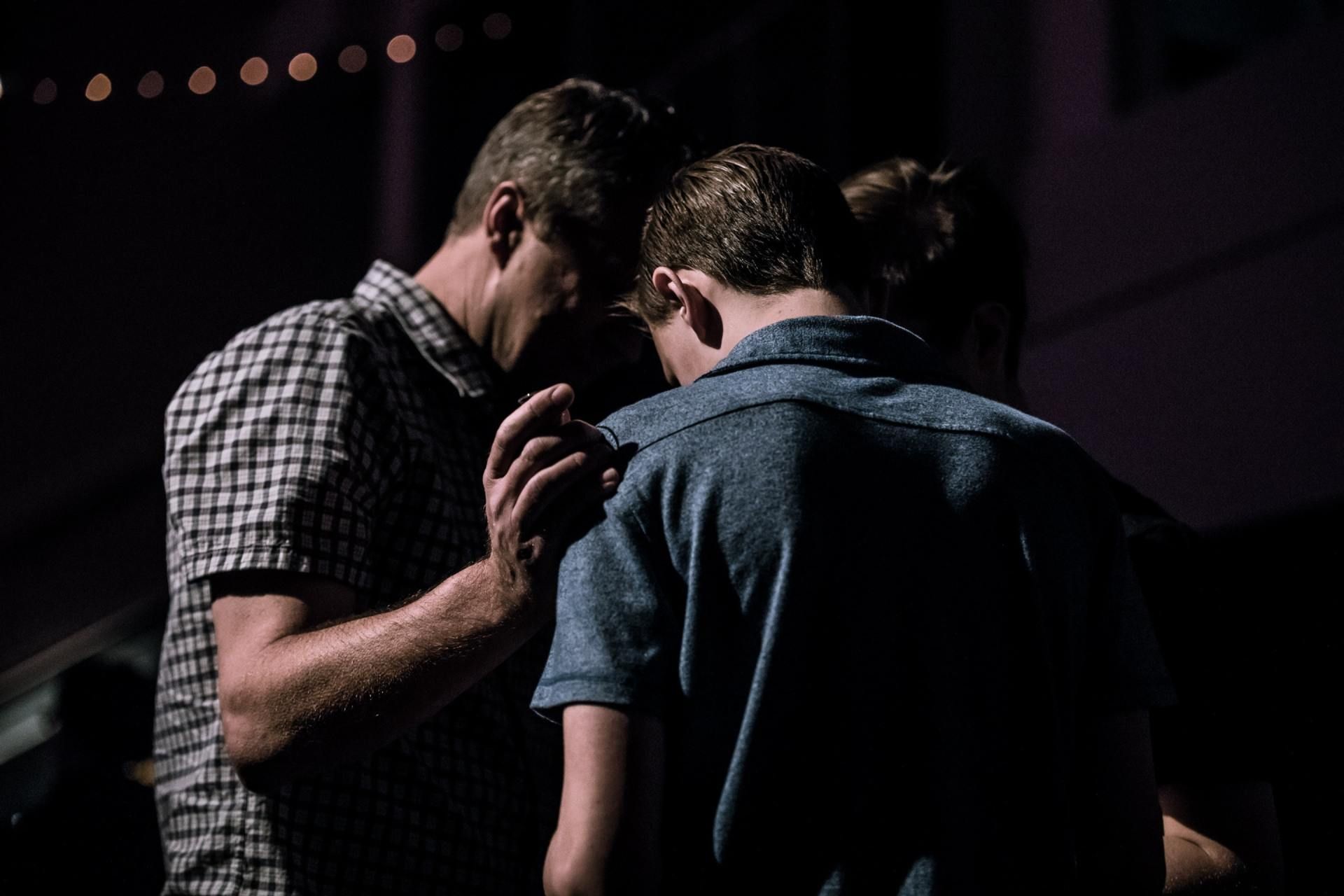 Three people with heads bowed, hands touching in prayer. Dark setting, soft light, focused on the figures.