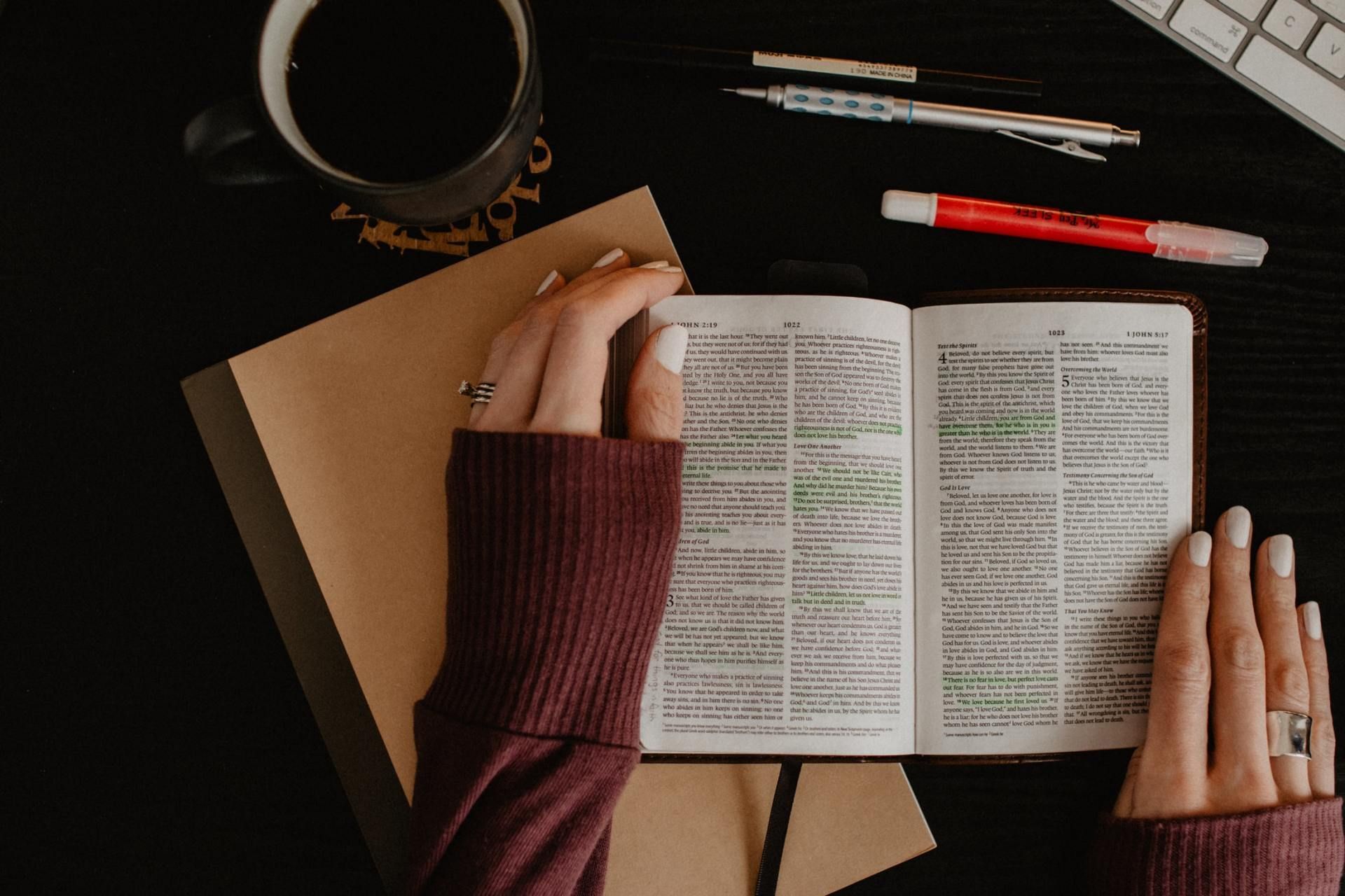 Hands holding an open Bible with green highlights, coffee, and pens on a black desk.