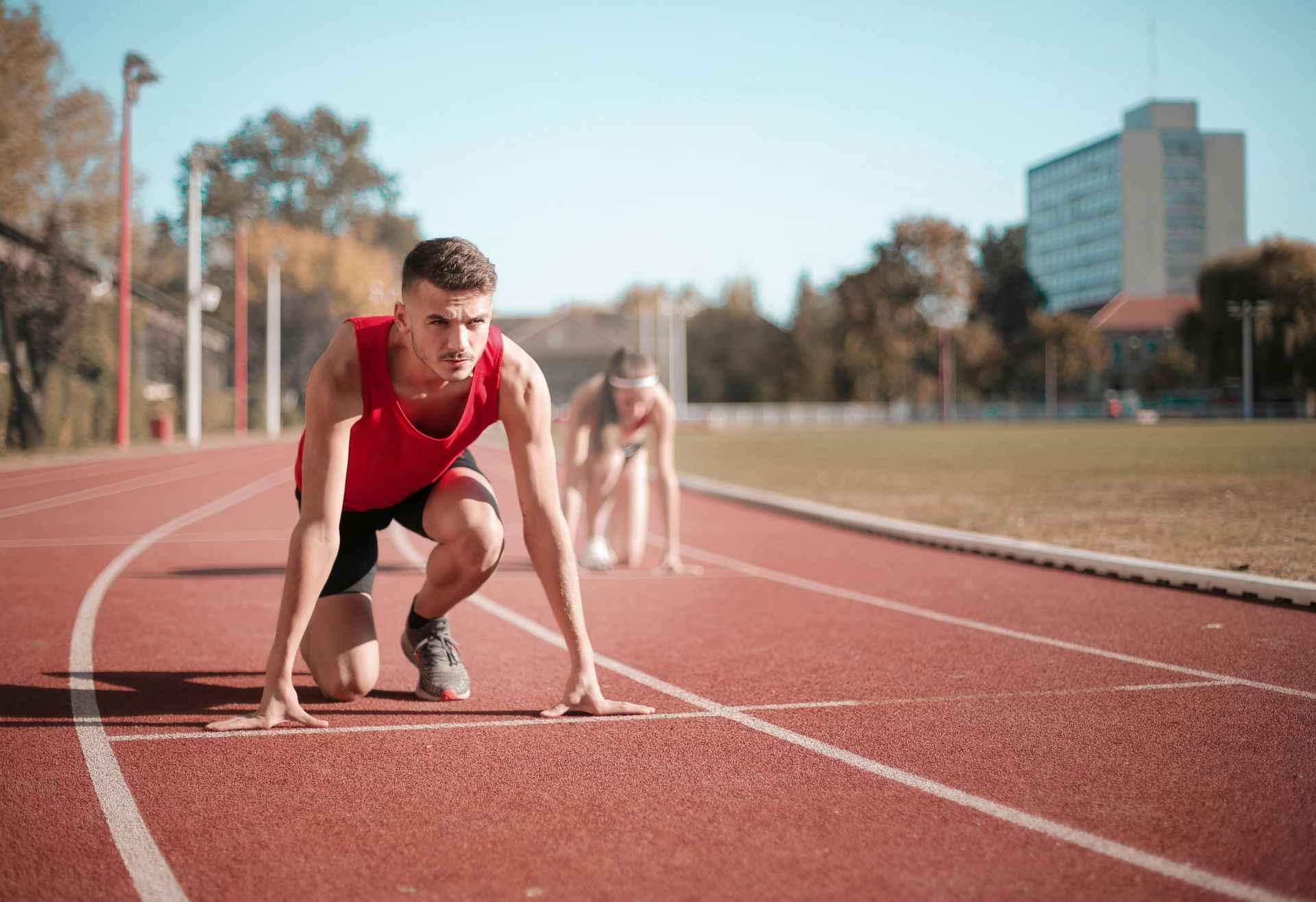 Man in red singlet at starting line, focused, on a track. Another athlete in the background.