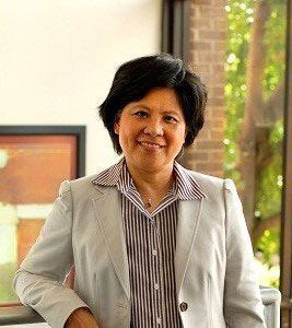 A woman in a suit and striped shirt is standing in front of a window and smiling.