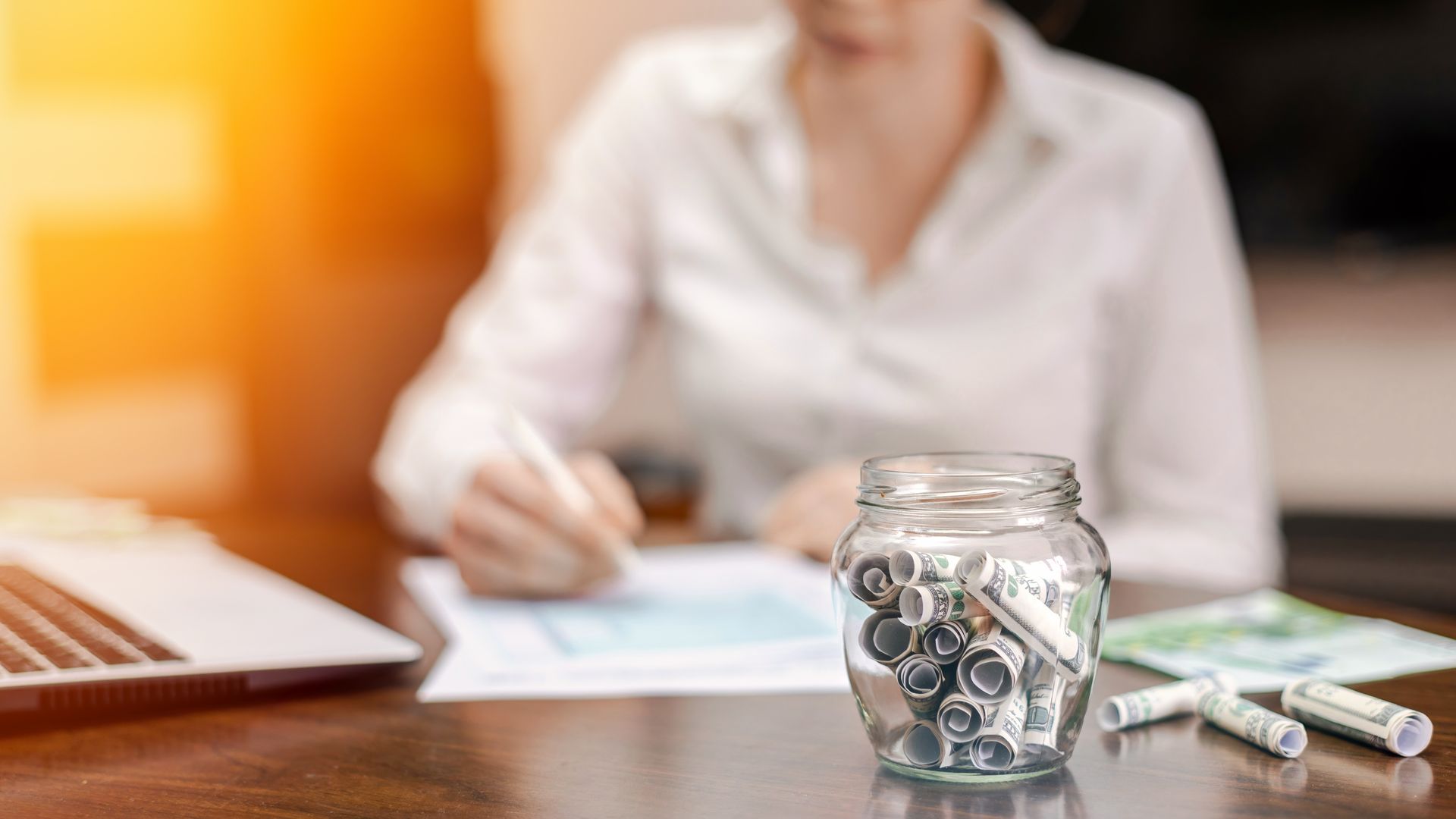 A woman is sitting at a desk with a jar of coins and a laptop.