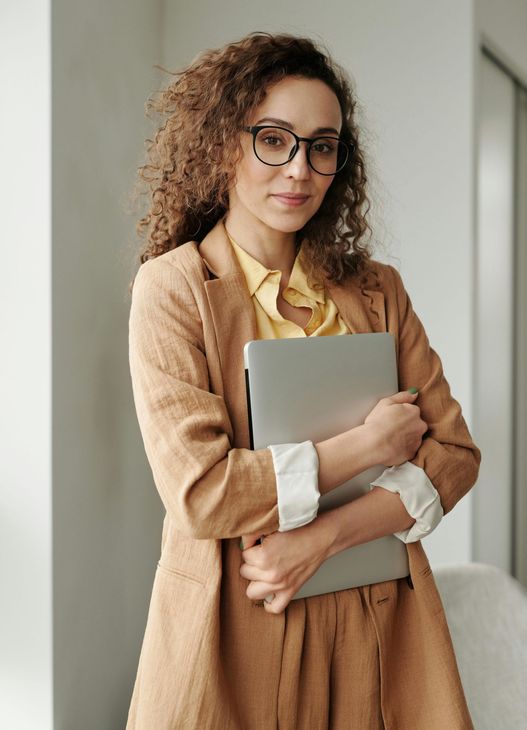 A woman wearing glasses is holding a laptop in her arms.