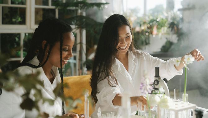 Two women are sitting at a table with a microscope and test tubes.