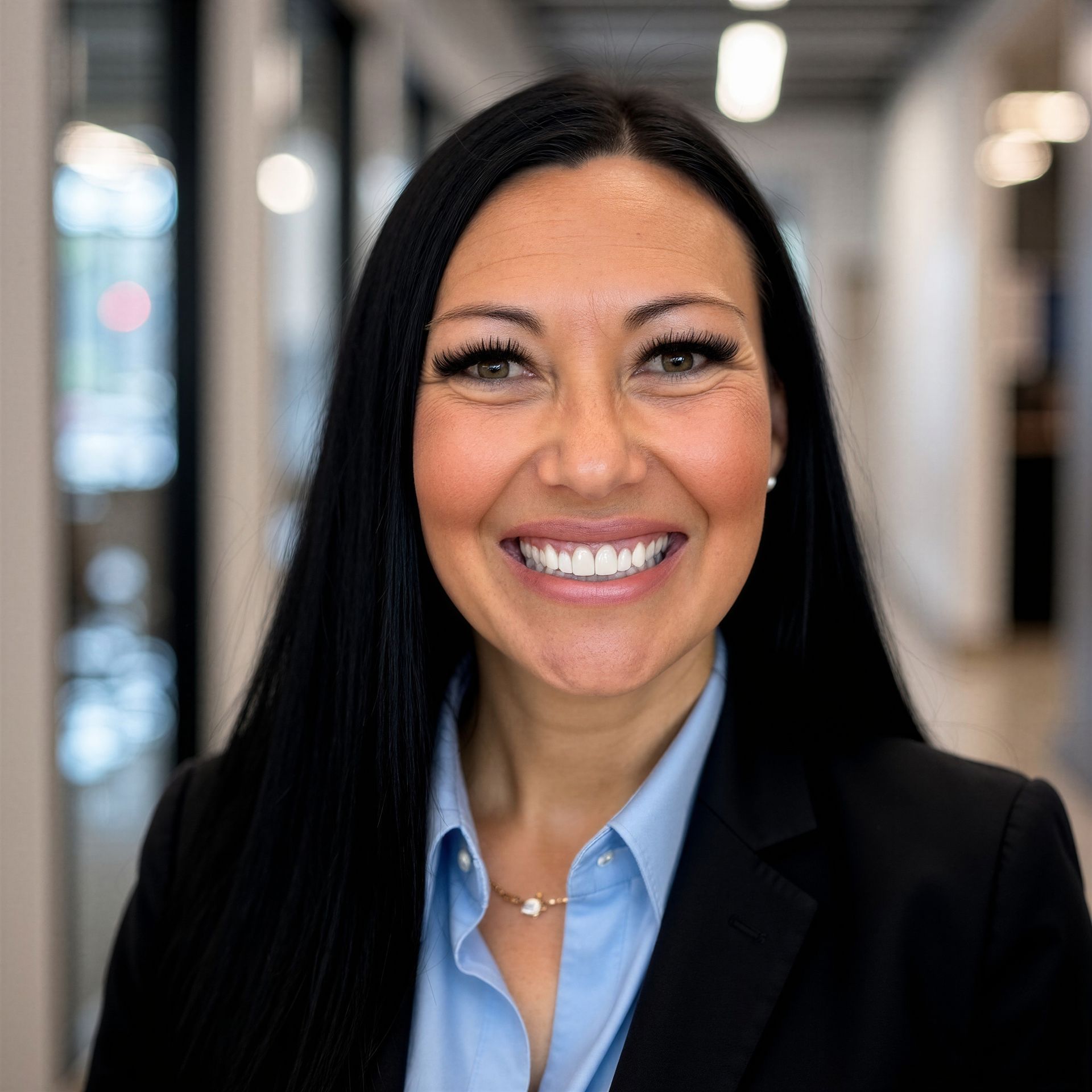 Smiling woman with long dark hair in black cardigan and white top, standing before a light wood wall.