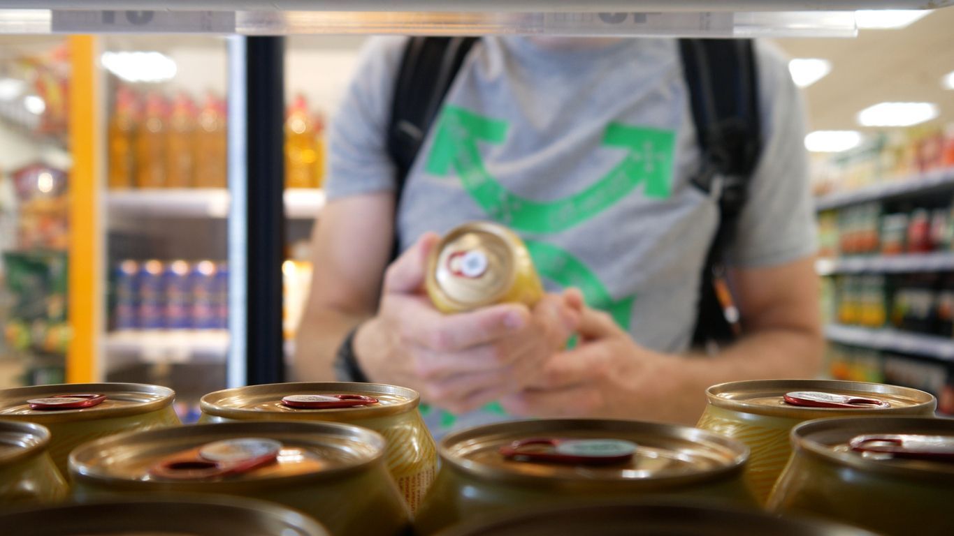 Person in a grocery aisle holding a can behind a row of canned goods in the foreground.
