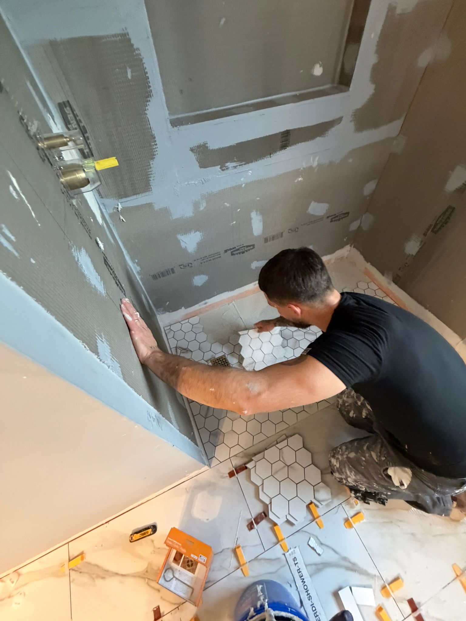 Man installing hexagon tiles in a bathroom, using leveling clips. Walls are unfinished drywall.