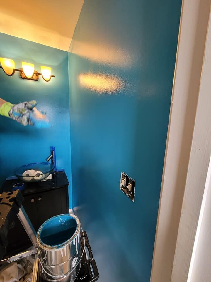 Person spray painting a bright blue bathroom wall. A paint can sits on a tray below.