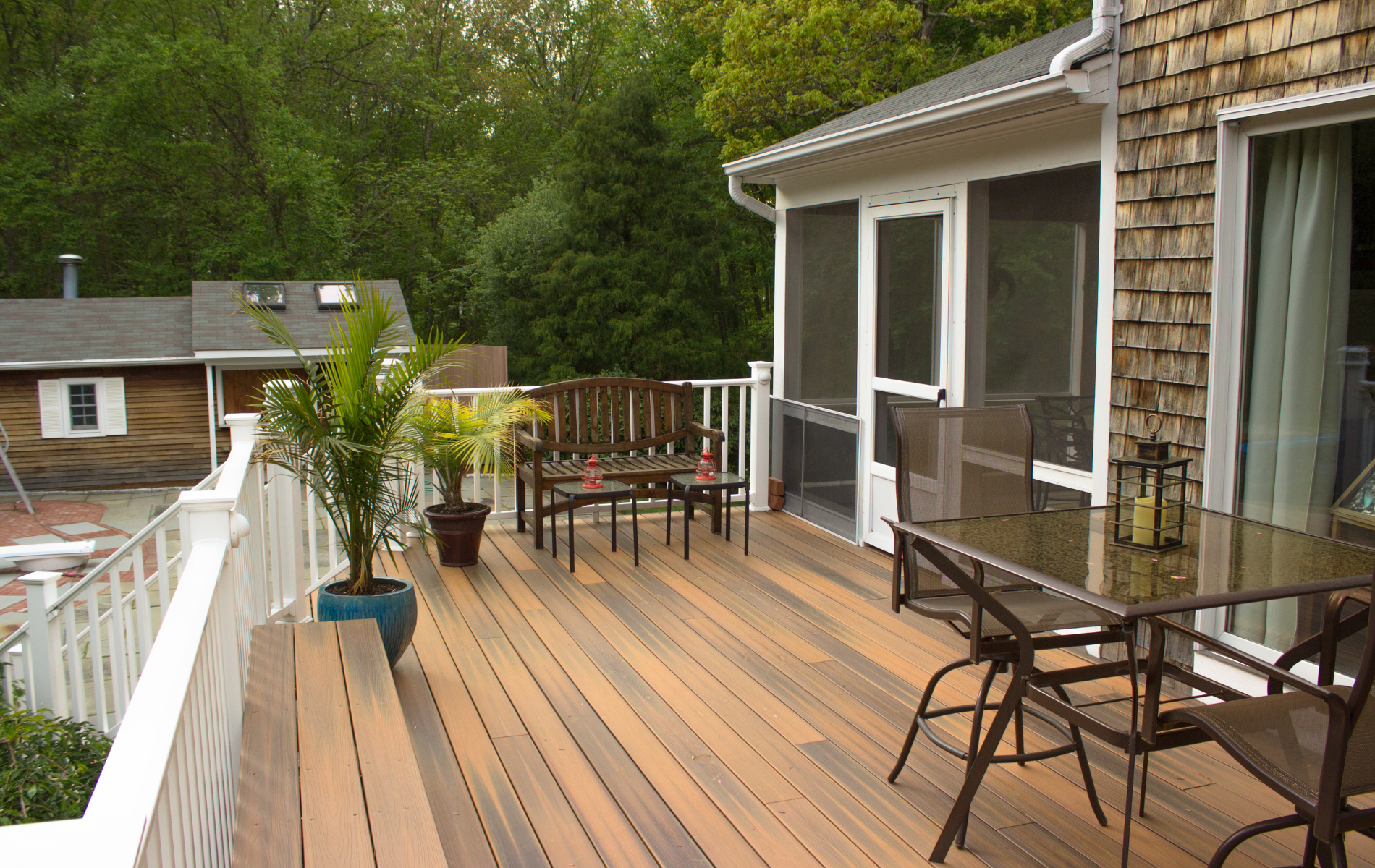 Wooden deck with outdoor furniture, potted plants, and screened porch. Overlooking a yard with trees.