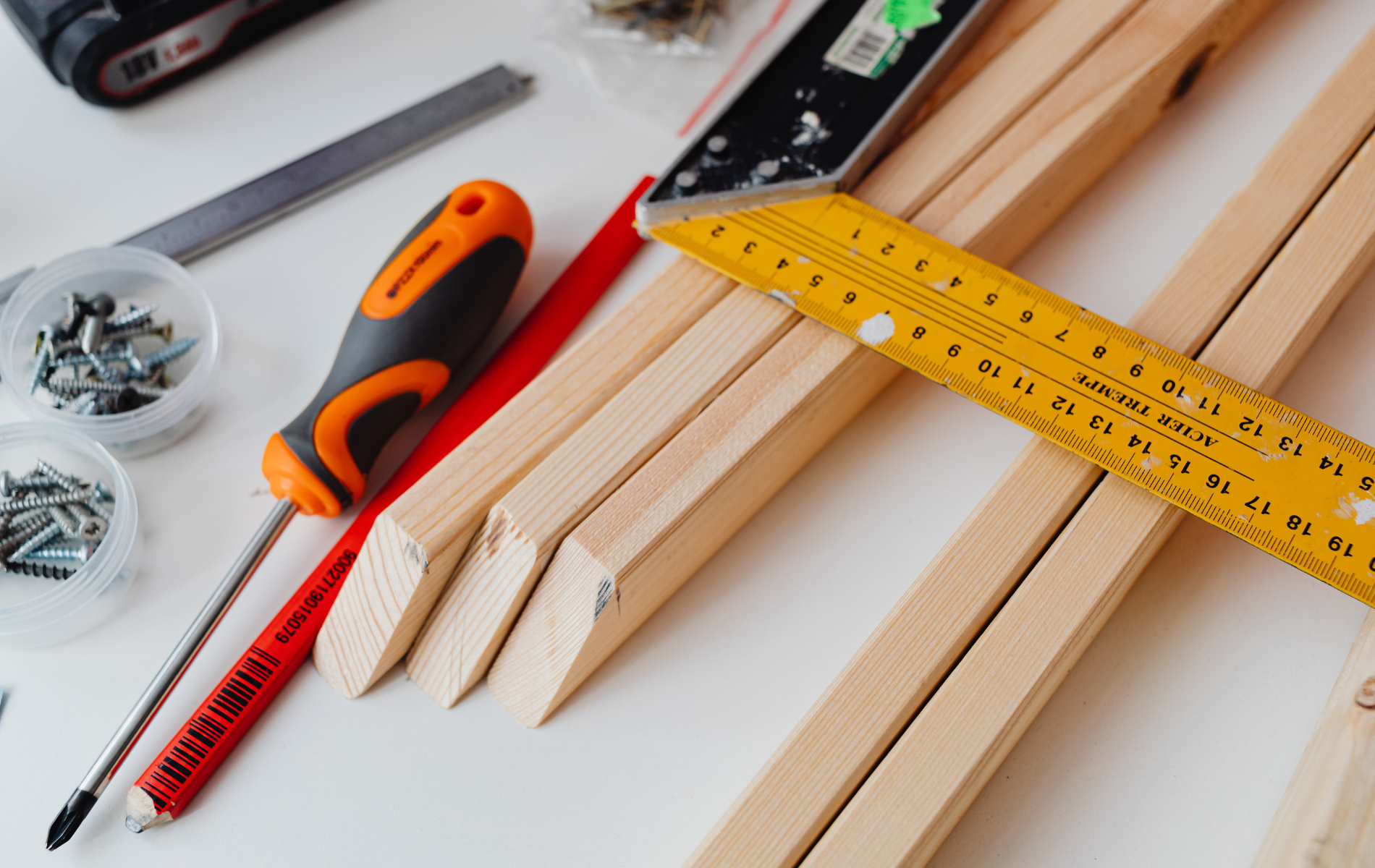 Wooden planks, screws, screwdriver, measuring tape, and pencil arranged on a white surface for a woodworking project.