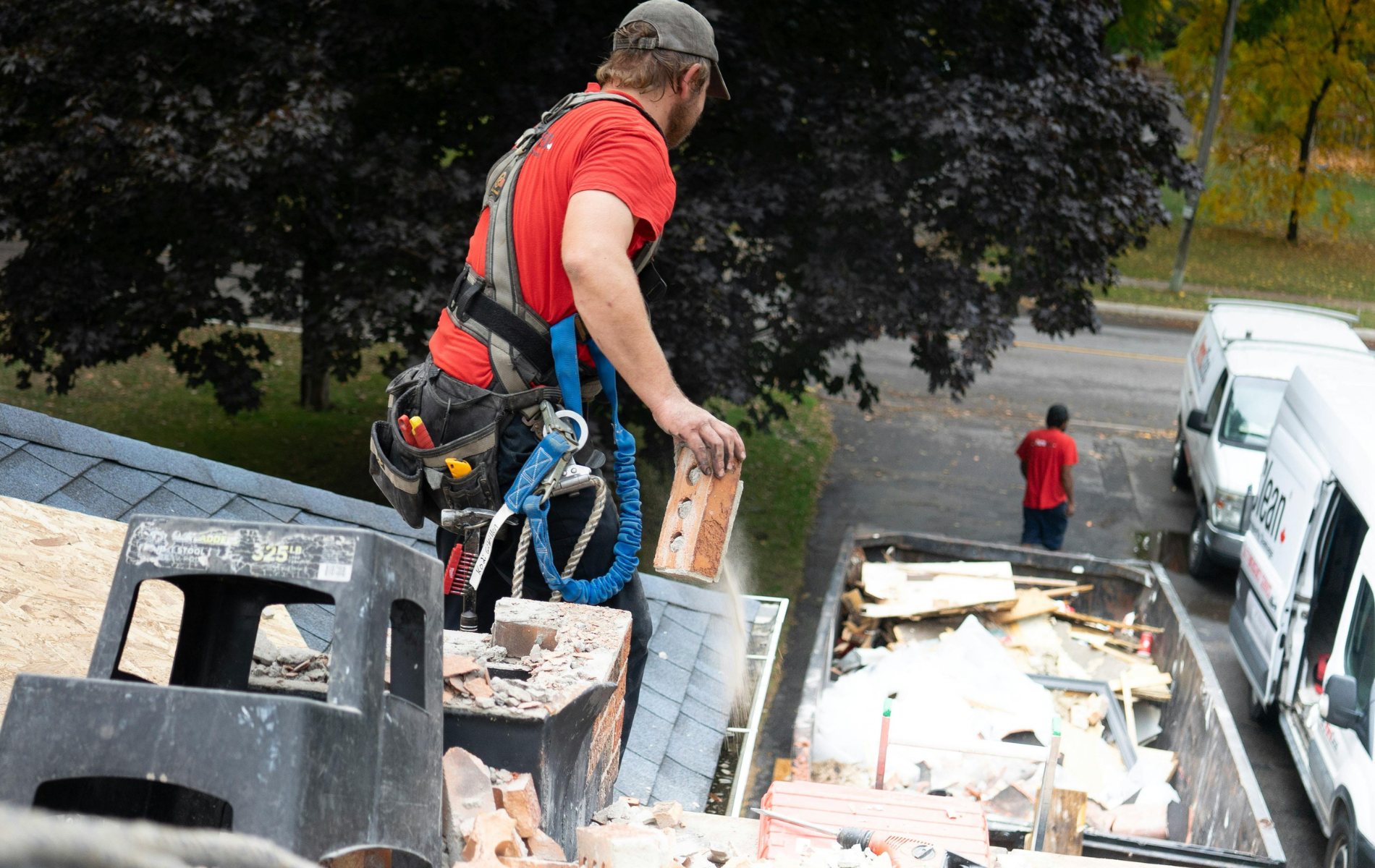 Roofer in safety harness removes bricks from a chimney and dumps them into a bin on a roof.