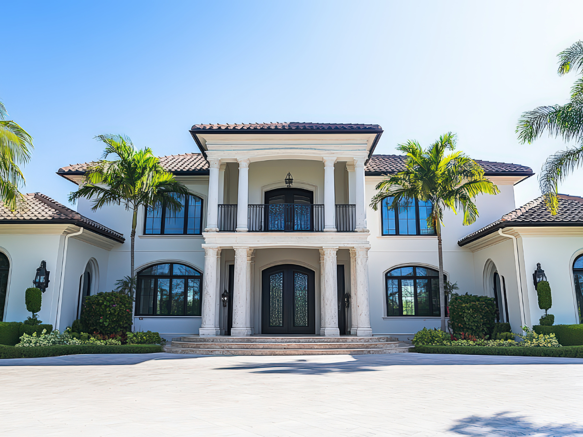 White mansion with columns, arched windows, and palm trees, driveway on a sunny day.
