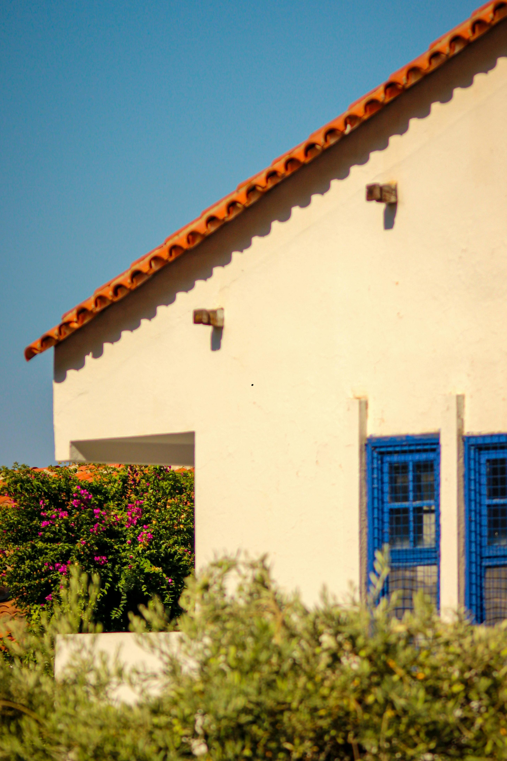 Off-white building with terracotta roof, blue window, and bougainvillea, under a clear blue sky.
