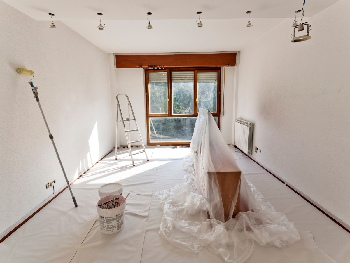 Room being painted, with white walls, drop cloths, ladder, and a paint roller.