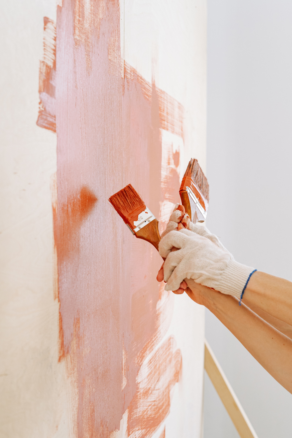 Hands wearing white gloves holding two paintbrushes, applying coral paint to a white wall.