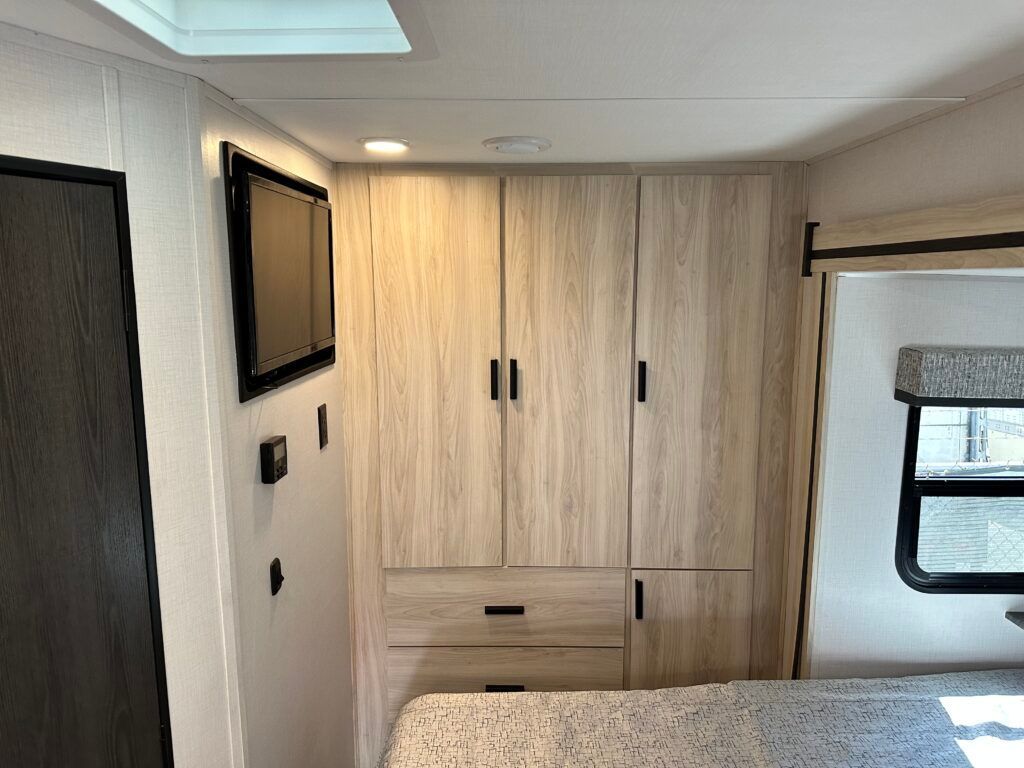 A bedroom in a rv with a bed , cabinets , drawers and a flat screen tv.