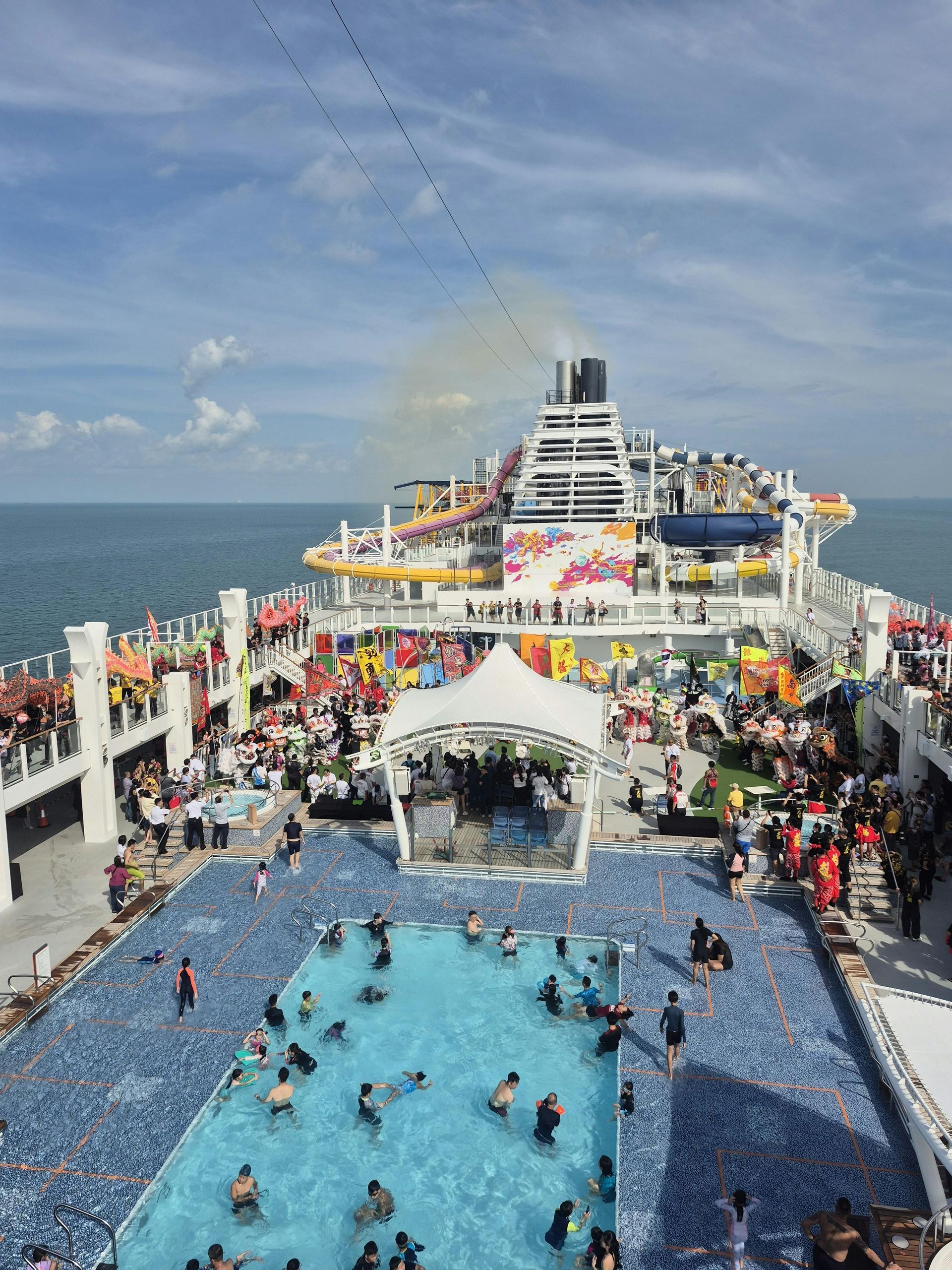Crowded cruise ship deck with pool, people swimming and lounging under a sunny blue sky