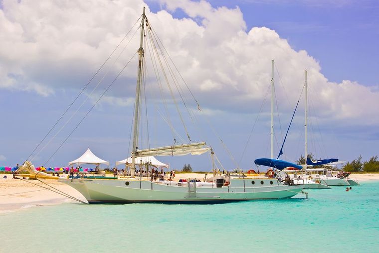 Sailboat anchored in turquoise water near a sandy beach under a cloudy sky, with tents and other boats nearby