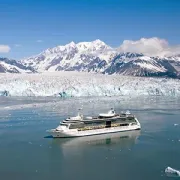 Cruise ship sailing past icy glacier and snow-covered mountains under a clear blue sky
