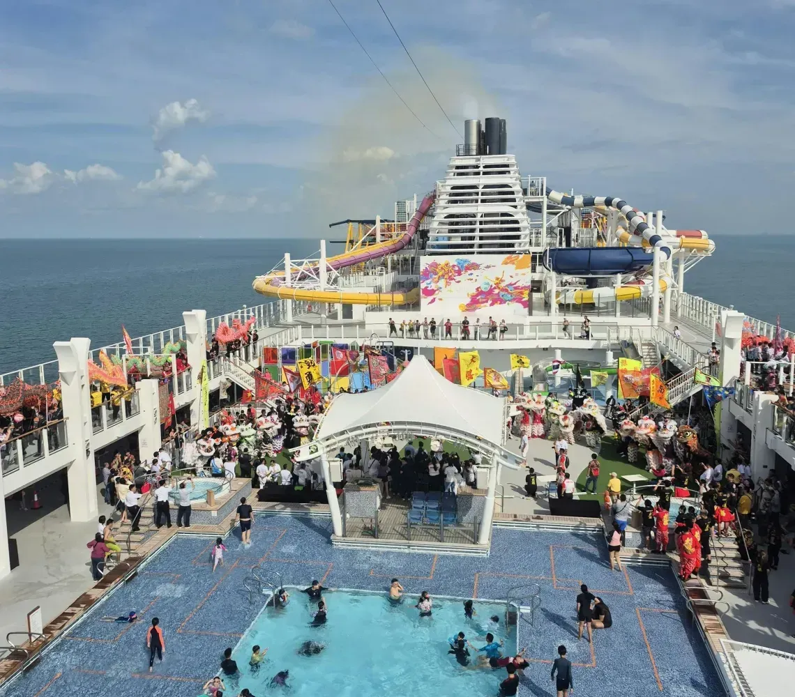Crowded cruise ship deck with pool, people swimming and lounging under a sunny blue sky