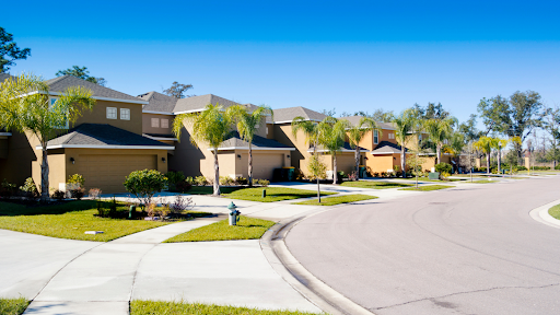 A beautiful suburban neighborhood and real estate with a row of modern, tan-colored houses featuring well-maintained lawns and palm trees.