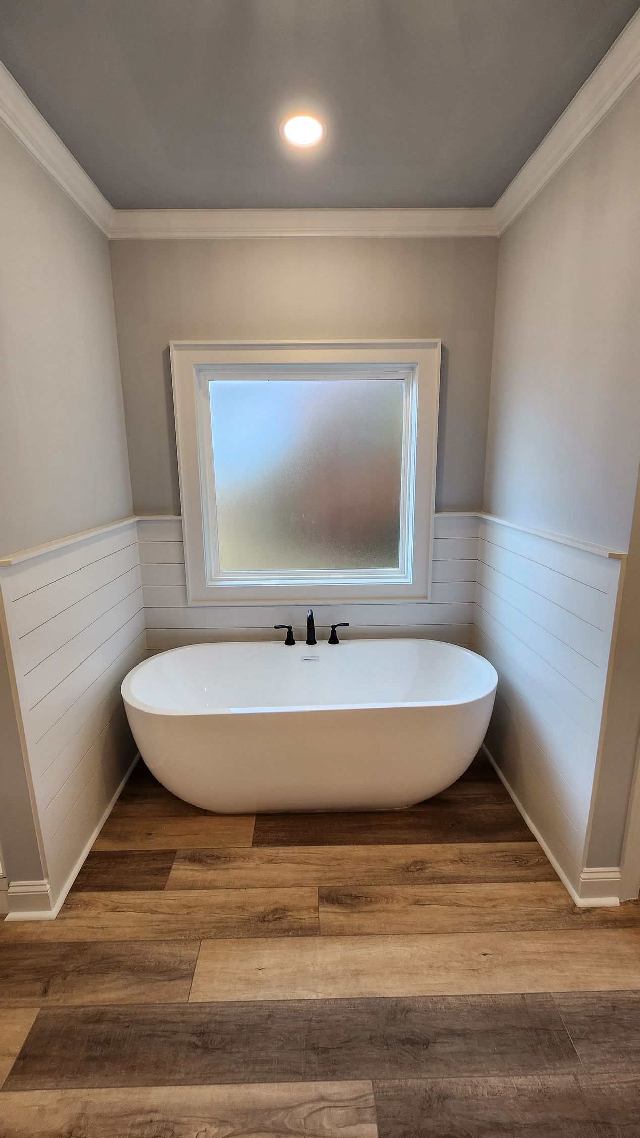 Freestanding white bathtub under a frosted window in a bathroom with wood-look flooring and light gray walls.