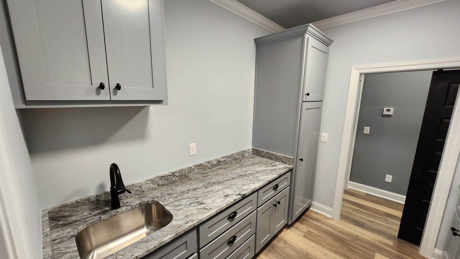 A utility room with grey cabinets, granite countertop, sink, and tall storage cabinet.  Doorway leads to another room.