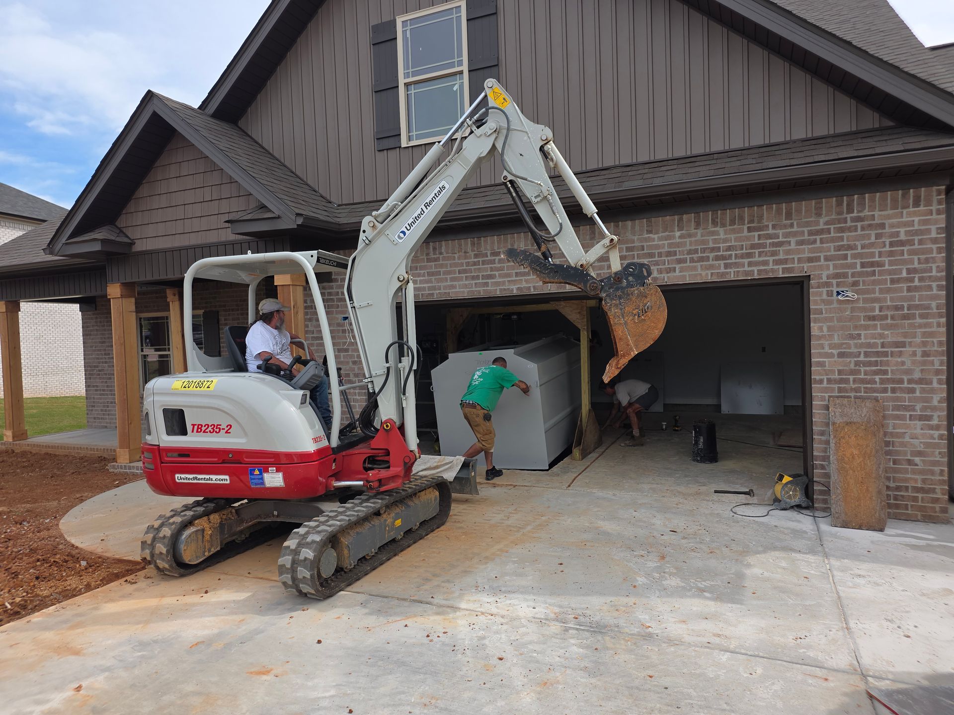 Excavator placing large object into a garage. Two people assist. House with brown siding in background.