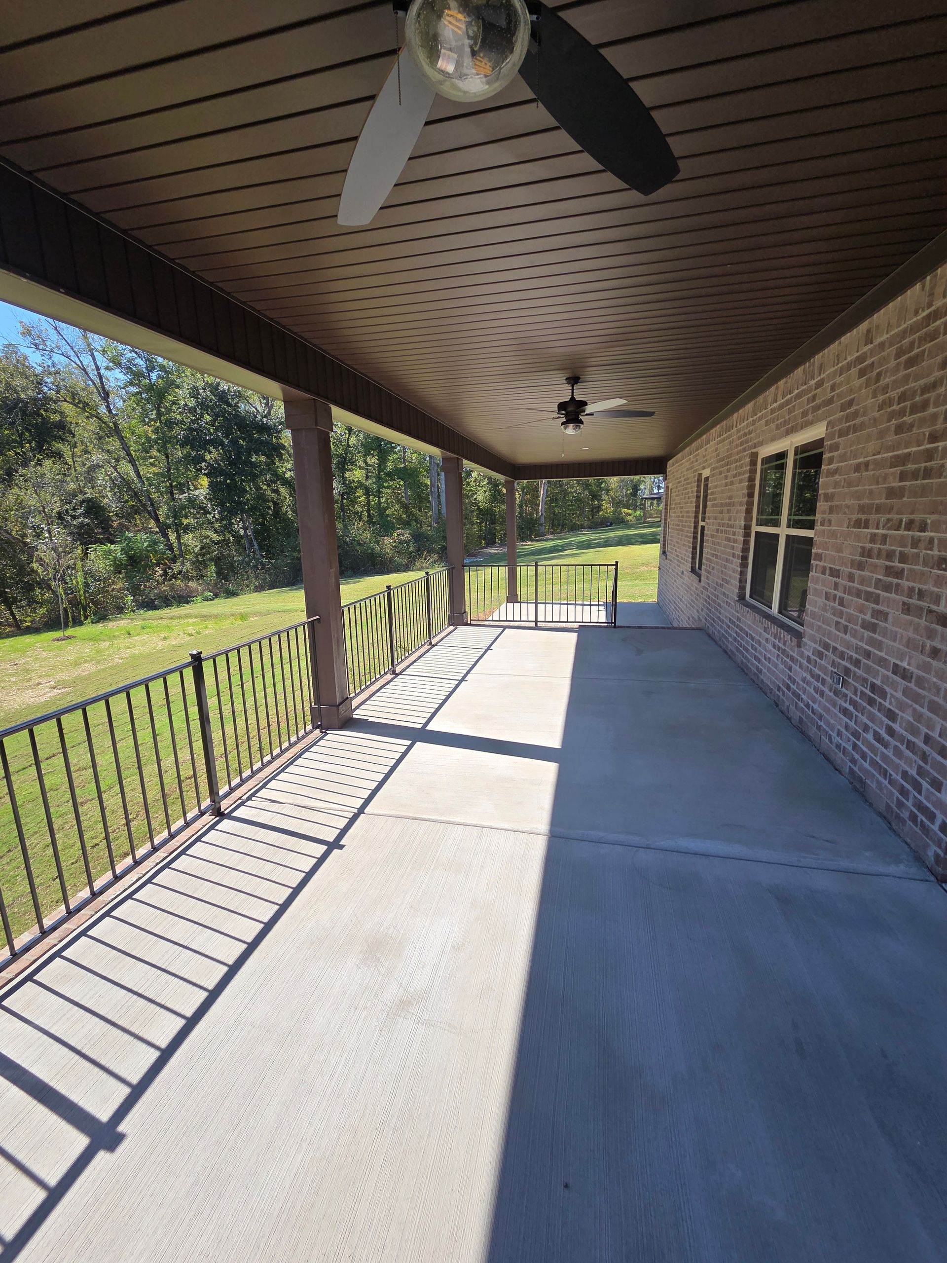 Covered patio with brick wall, concrete floor, ceiling fans, and black railing overlooking grass and trees.