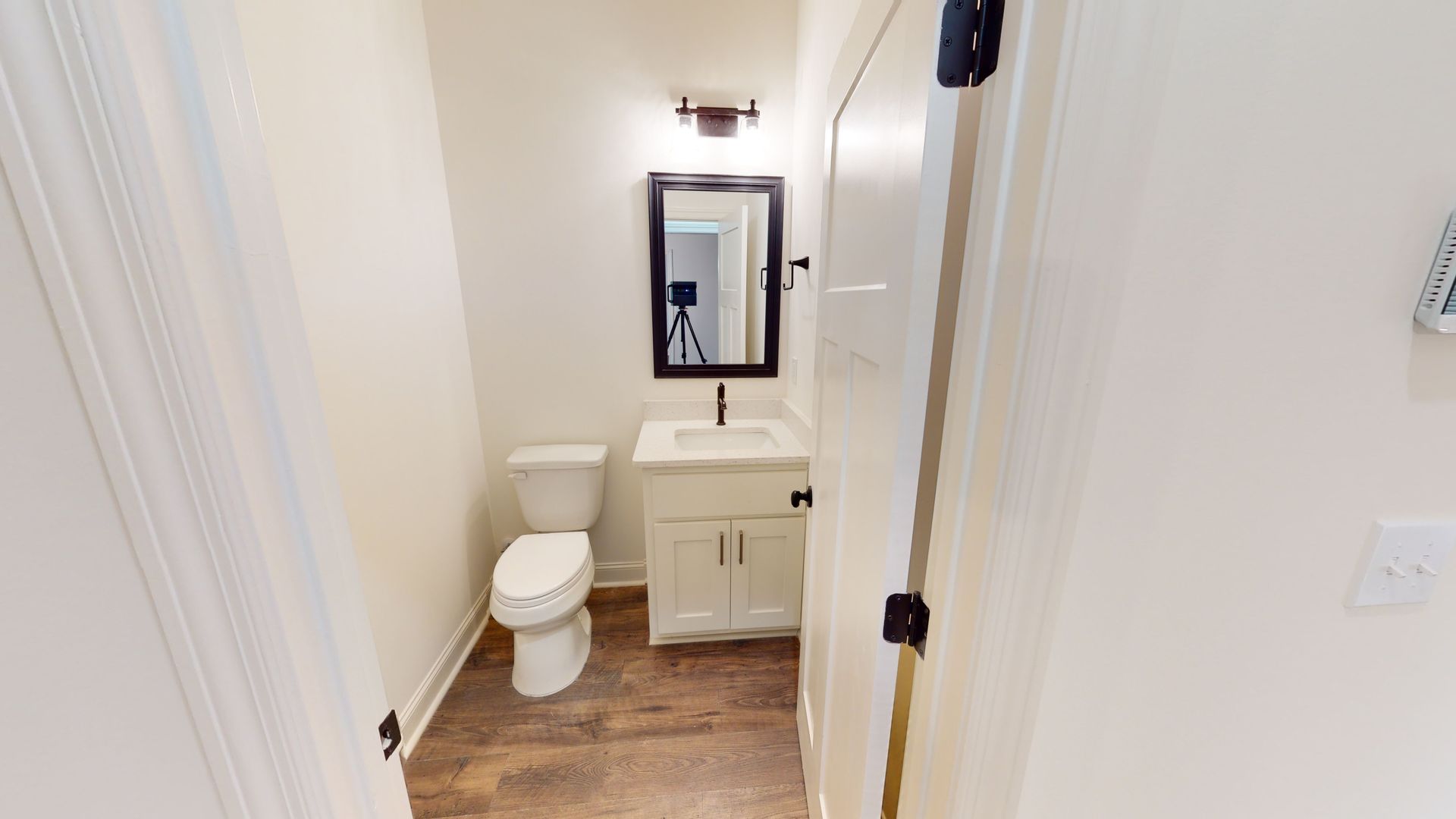 Small bathroom with a toilet, sink, and mirror. White walls and wooden floor.