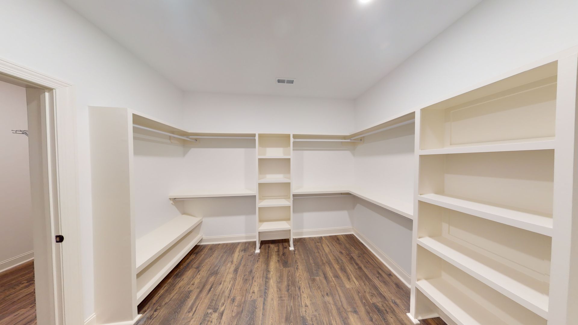 Empty white walk-in closet with shelves, rods, and wood-look flooring. A door is on the left.