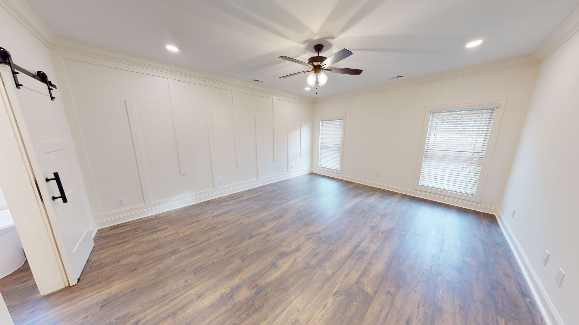 Empty bedroom with wood-look flooring, white walls, two windows, barn door, and a ceiling fan.