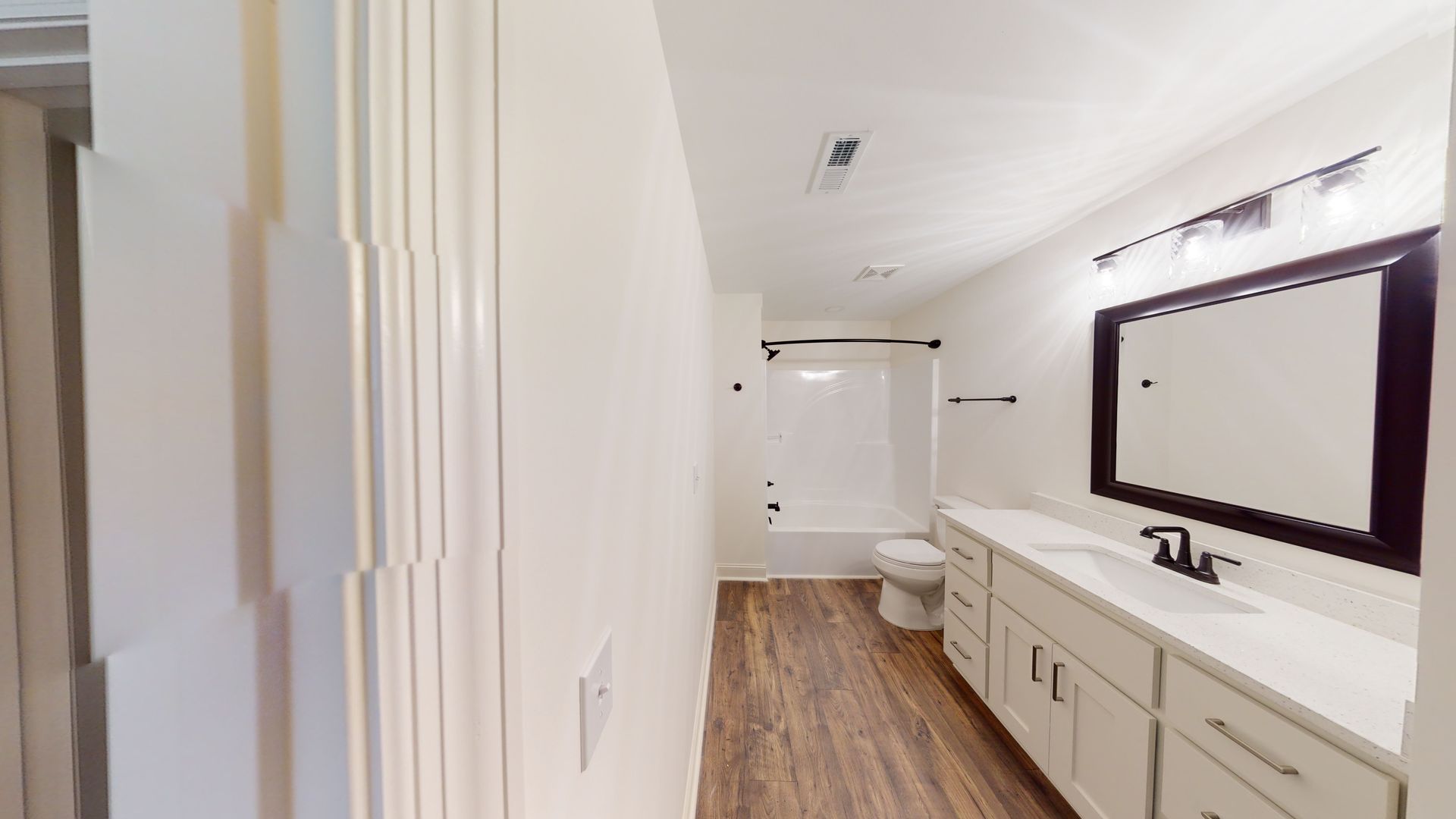 Bathroom with white walls, vanity, and shower. Dark framed mirror and brown floor.