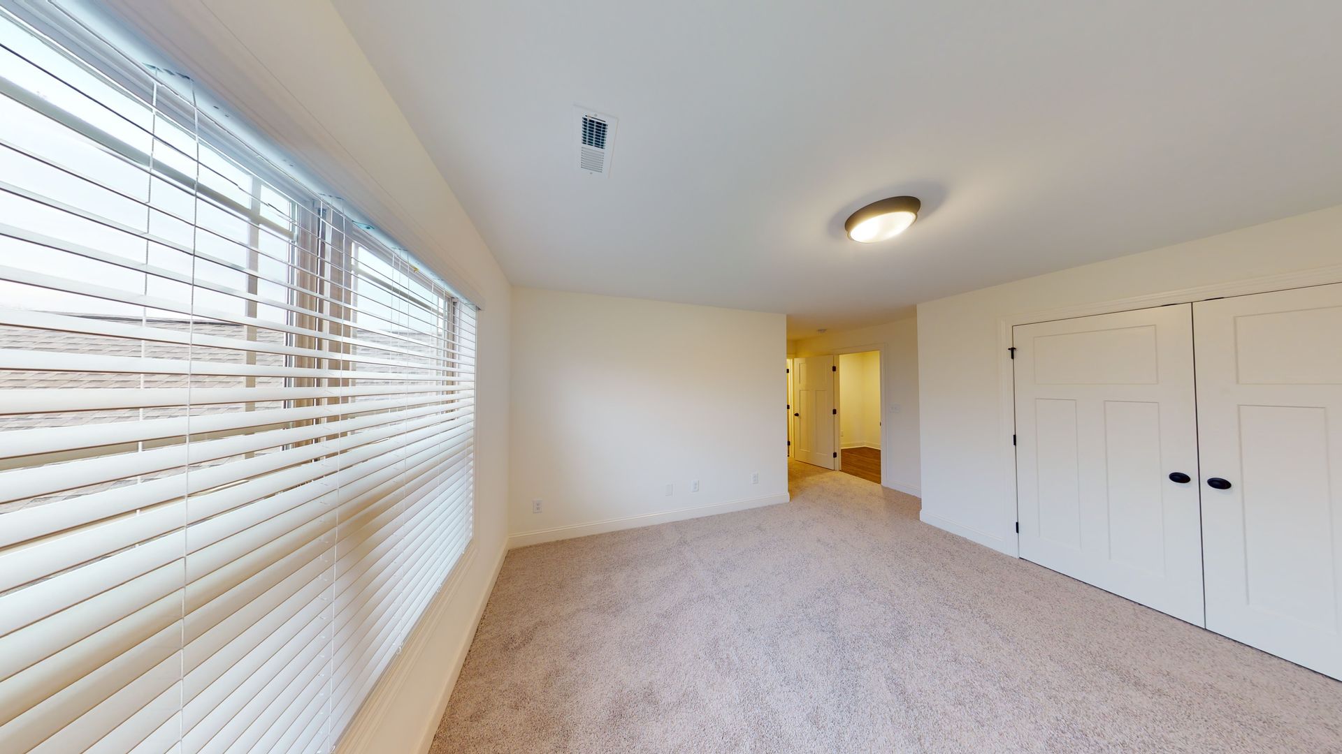 Empty room with beige carpet, window with blinds, and two white closet doors.
