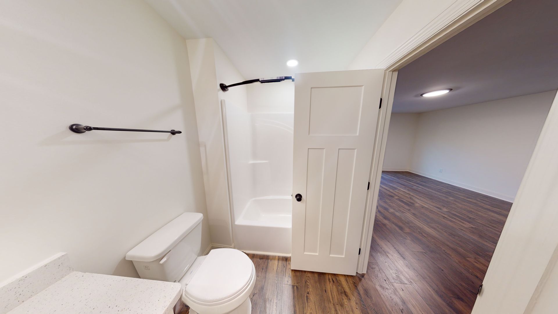 Bathroom with white walls, toilet, shower, and open door leading to a room with wood flooring.