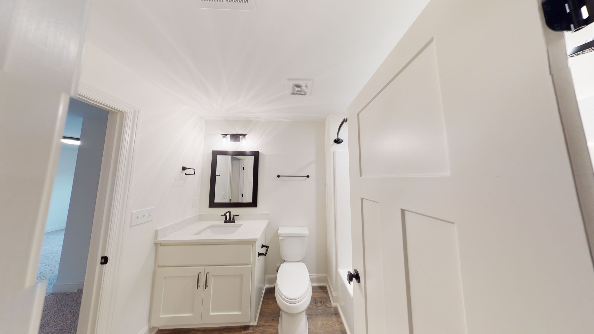 White bathroom with a vanity, toilet, and mirror.  Door on the left leads to another room.