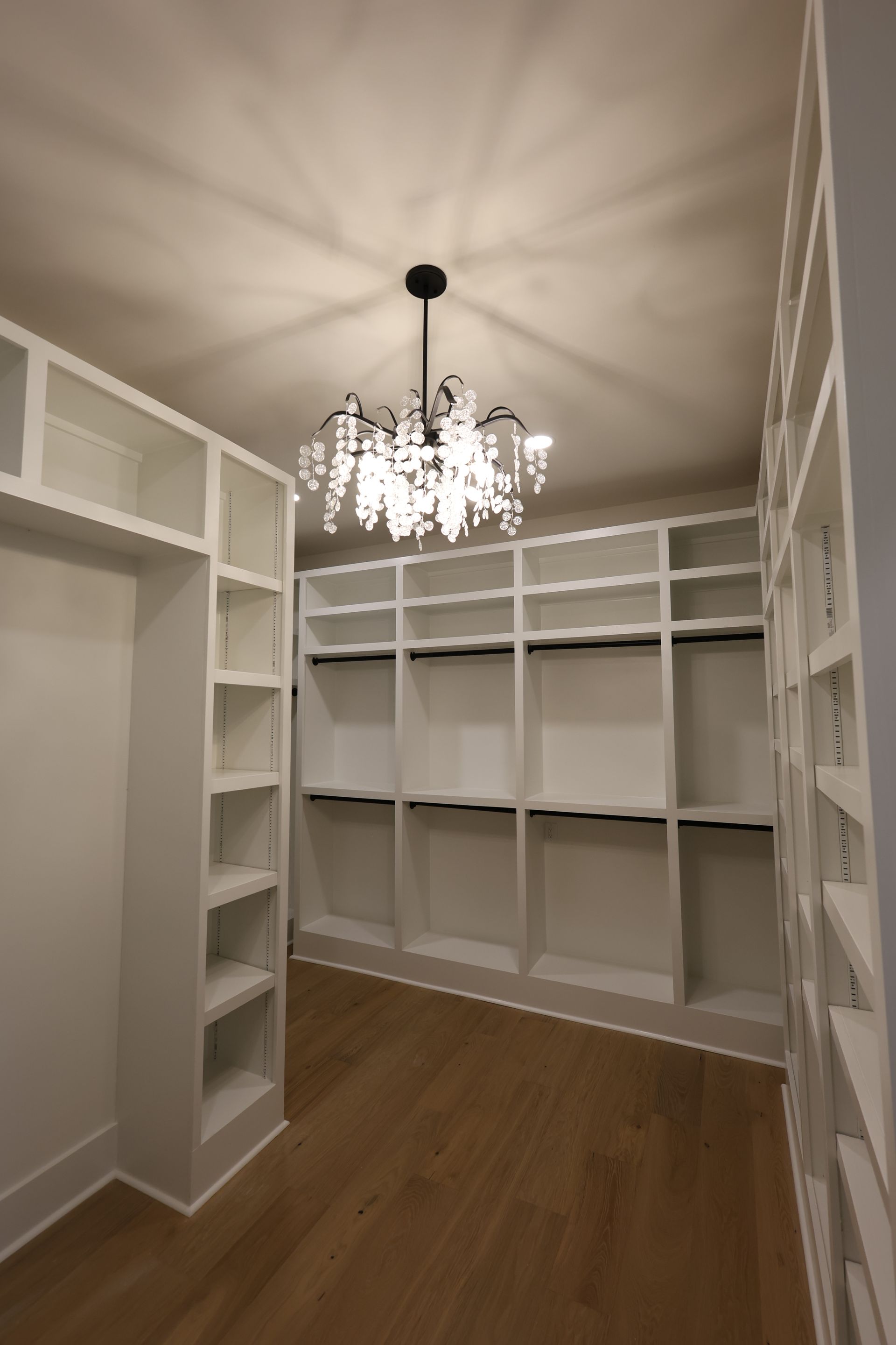 Empty white walk-in closet with shelves and rods, hardwood floor, chandelier lighting.