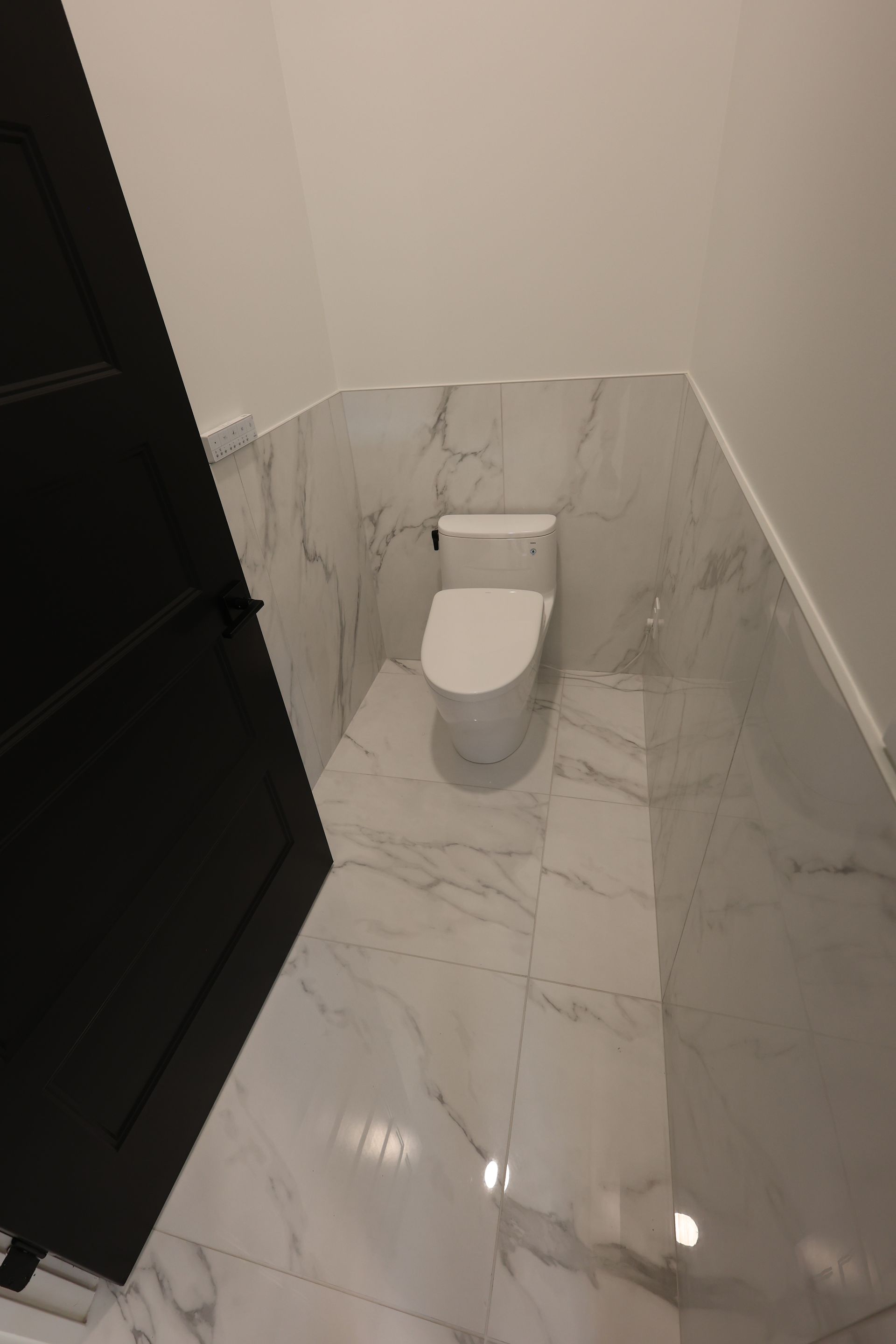A small, modern bathroom with a white toilet, marble-like tiled walls and floor, and a black door on the left.