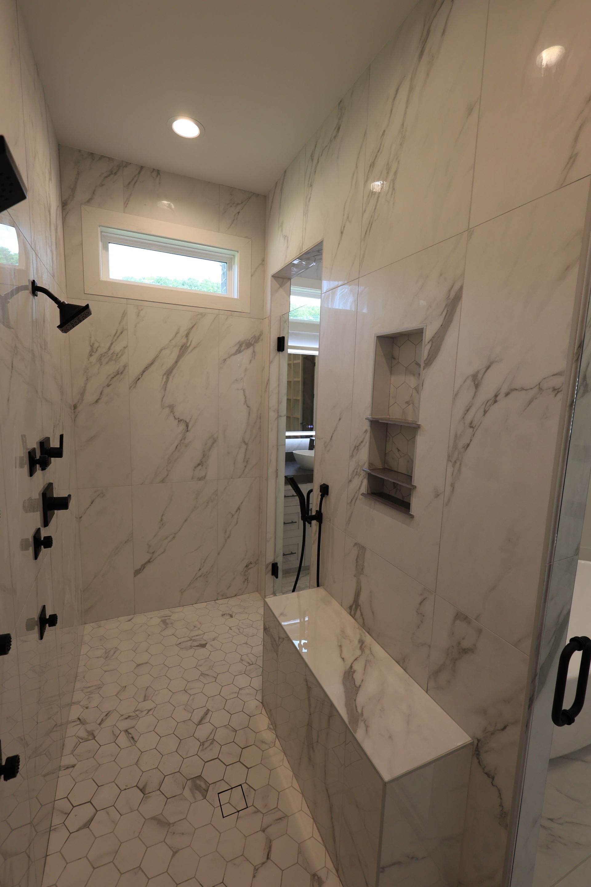 Spacious marble-tiled shower with a bench, black fixtures, and a glass door.