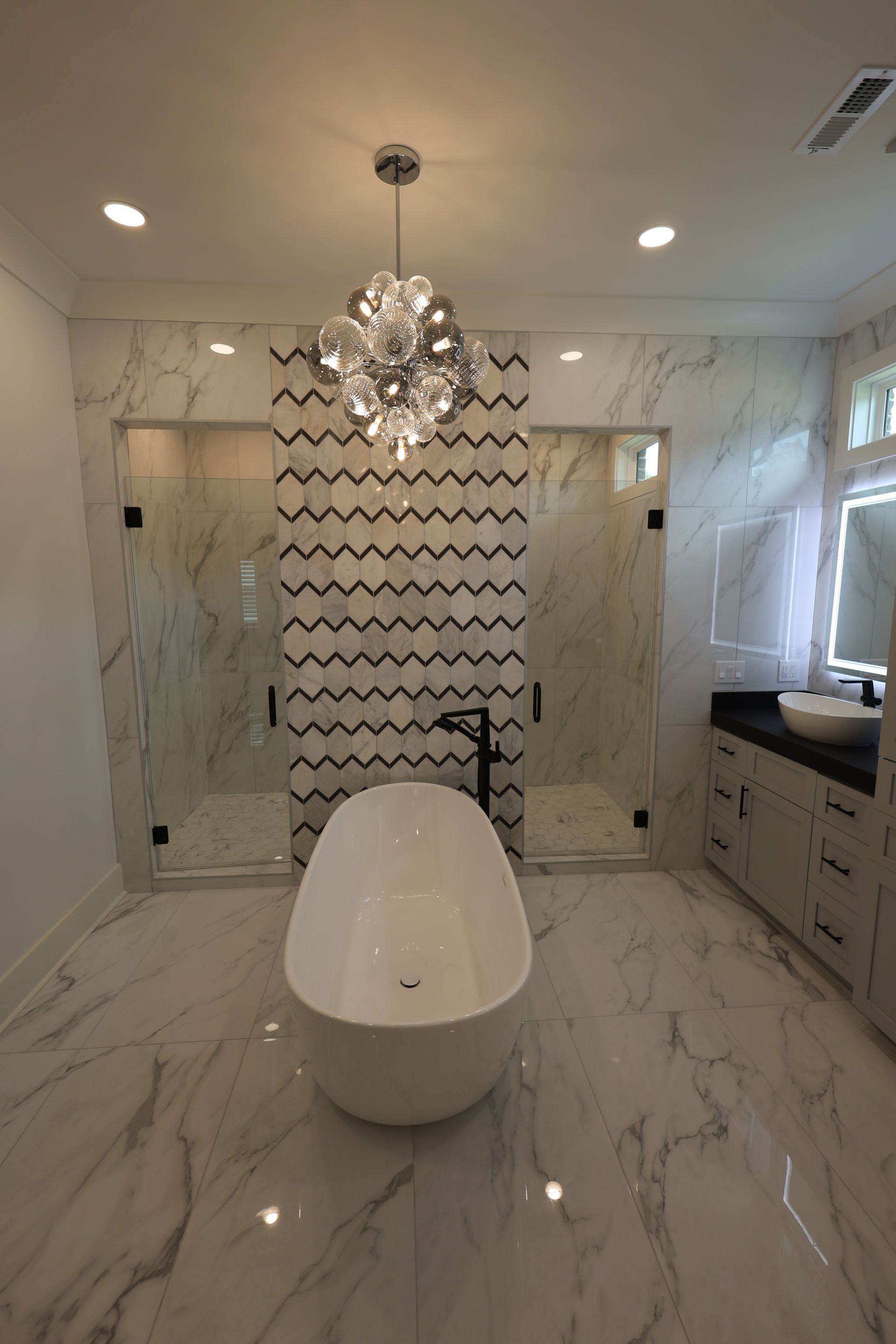 Luxurious bathroom with freestanding tub, two showers, chandelier, marble tile, and vanity.