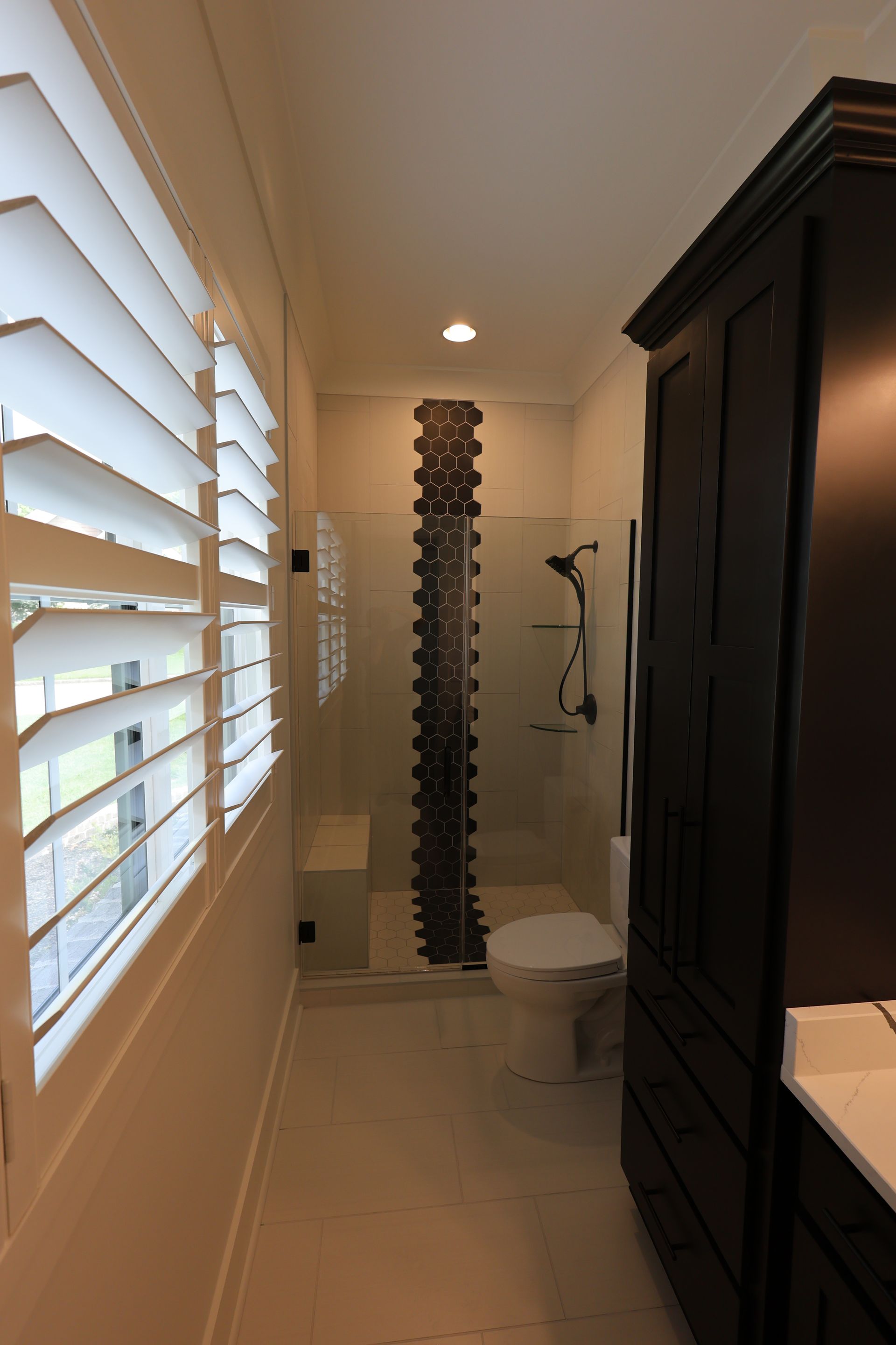 Narrow bathroom with white shutters, dark cabinet, glass shower, and toilet.