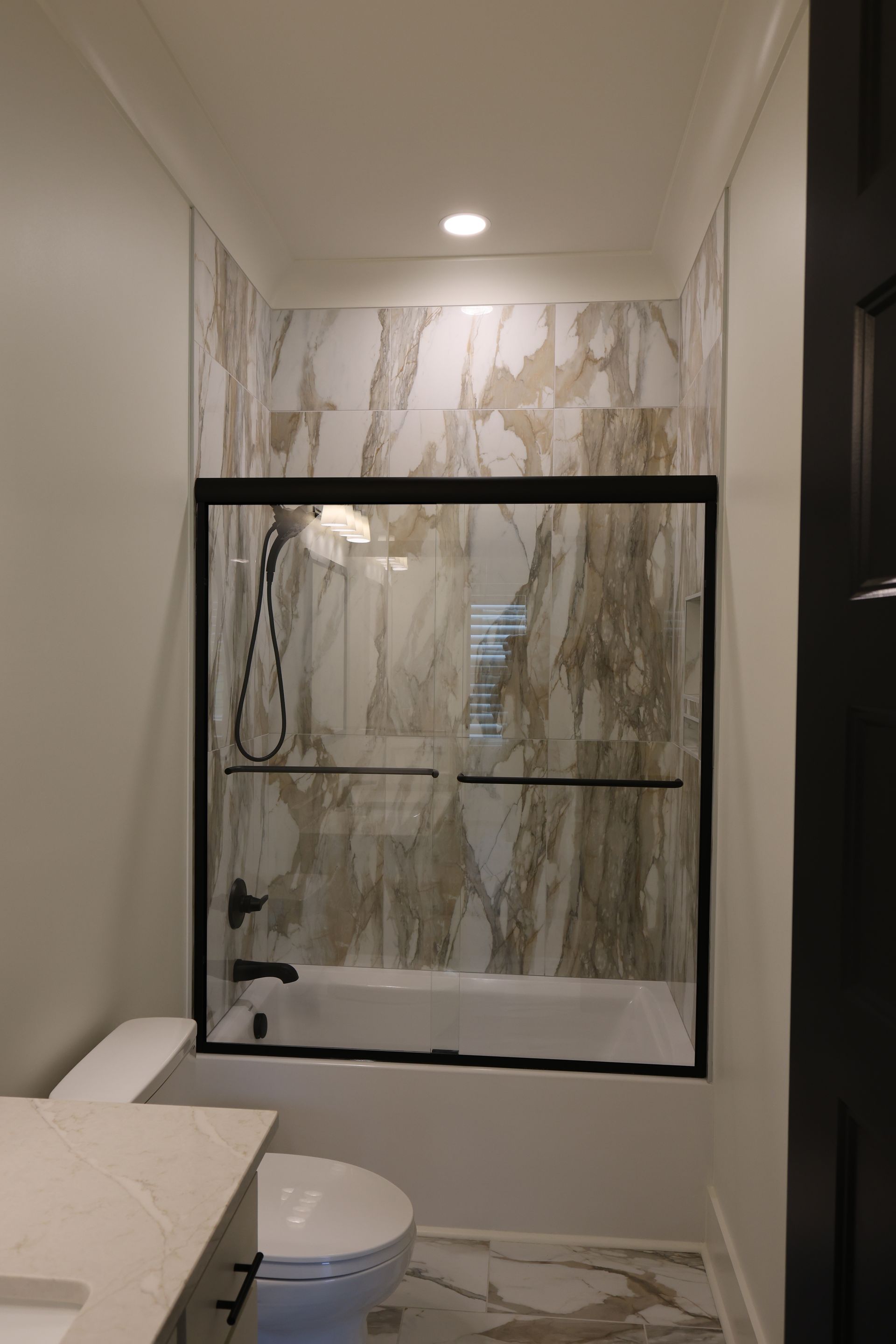 Bathroom with marble-look tile, black framed shower door, white tub, and toilet.