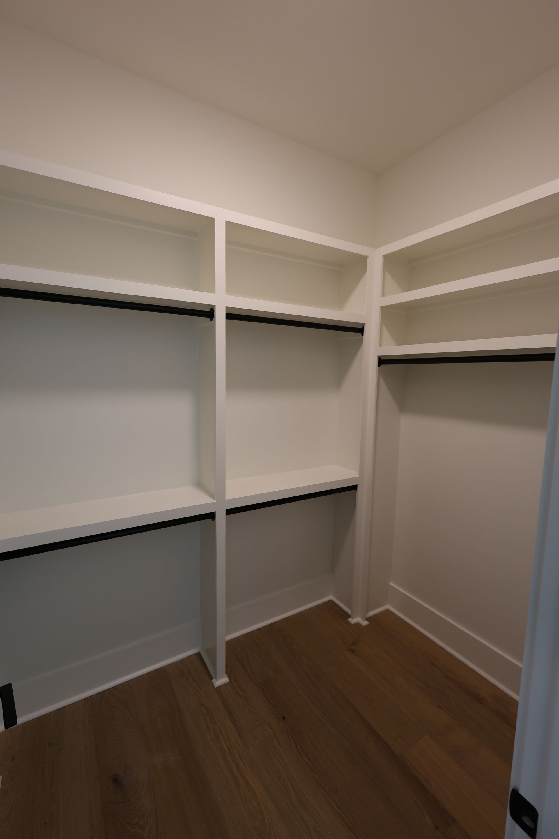 White closet with shelving and black rods, empty, with wood floor.