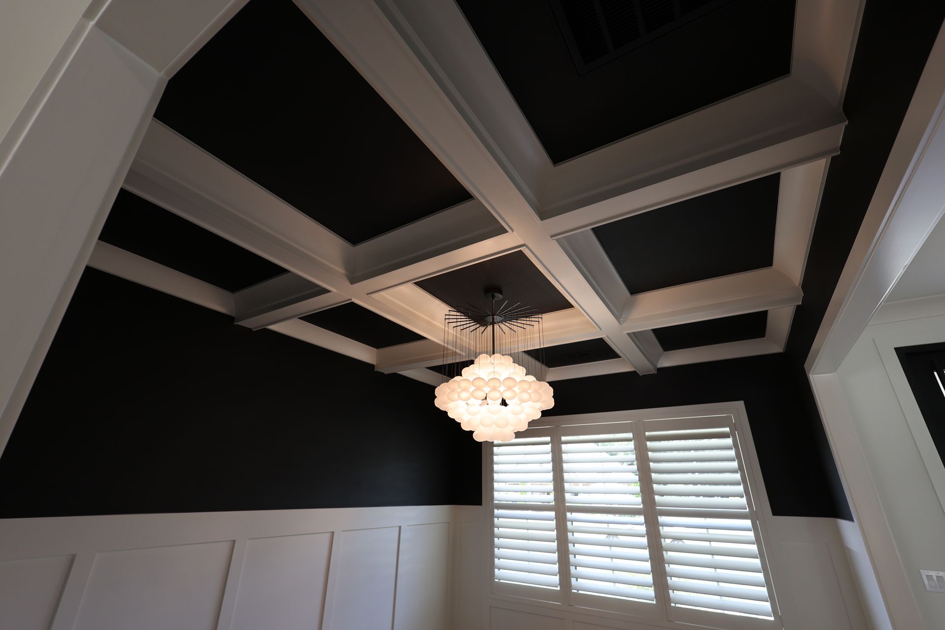 Black and white coffered ceiling with a chandelier in a room with a window and shutters.